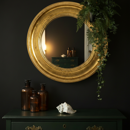 Round gold gilt mirror styled against a dark moody wall with green vintage chest of drawers and brown apothecary bottles