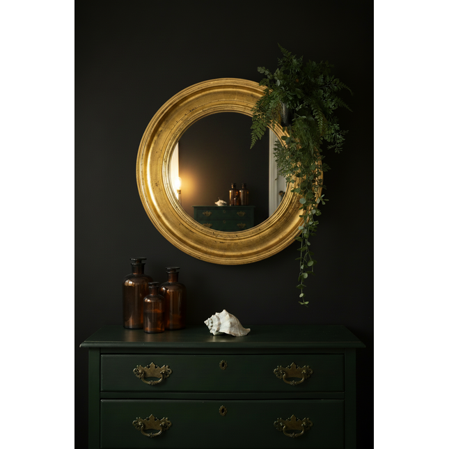 Round gold gilt mirror styled against a dark moody wall with green vintage chest of drawers and brown apothecary bottles