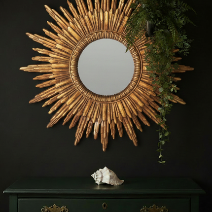 Gold sunburst wall mirror styled against a dark moody wall with green vintage chest of drawers and trailing foliage