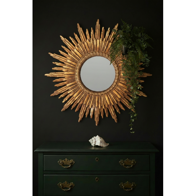 Gold sunburst wall mirror styled against a dark moody wall with green vintage chest of drawers and trailing foliage